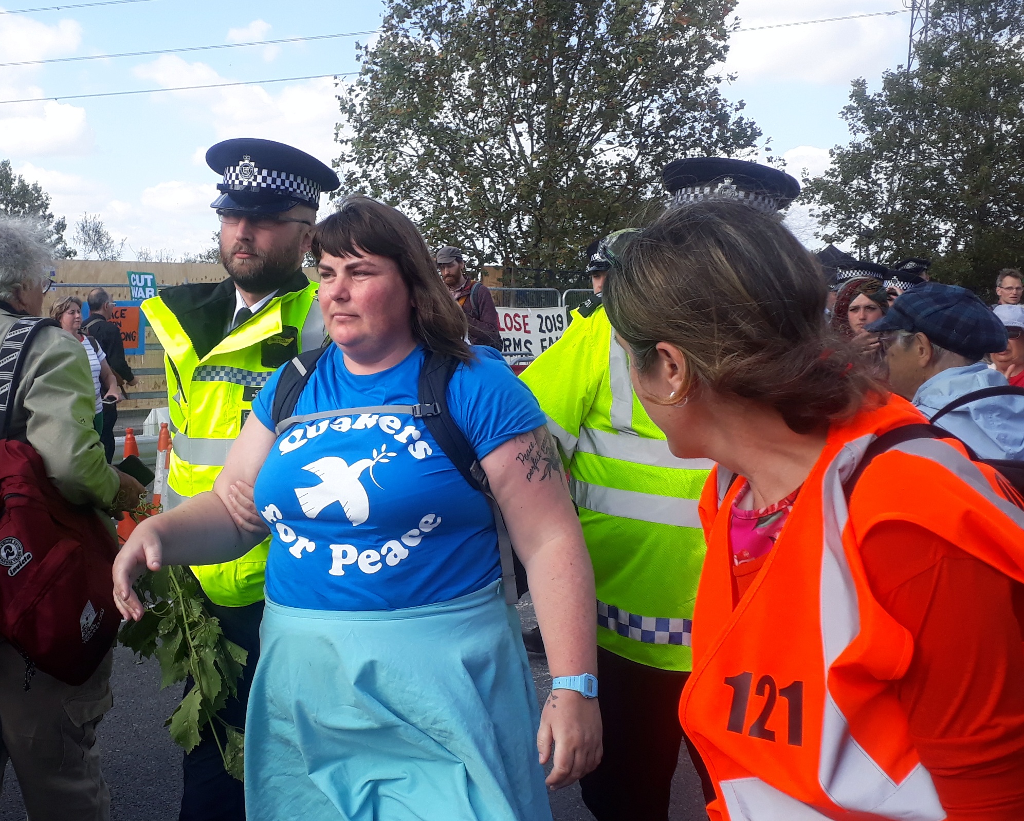 An activist with a 'Quakers for peace' t-shirt is held by the arms by two police officers on either side following her arrest on the No Faith in War day, 2019. A legal observer with a high-vis jacked on is talking to her.
